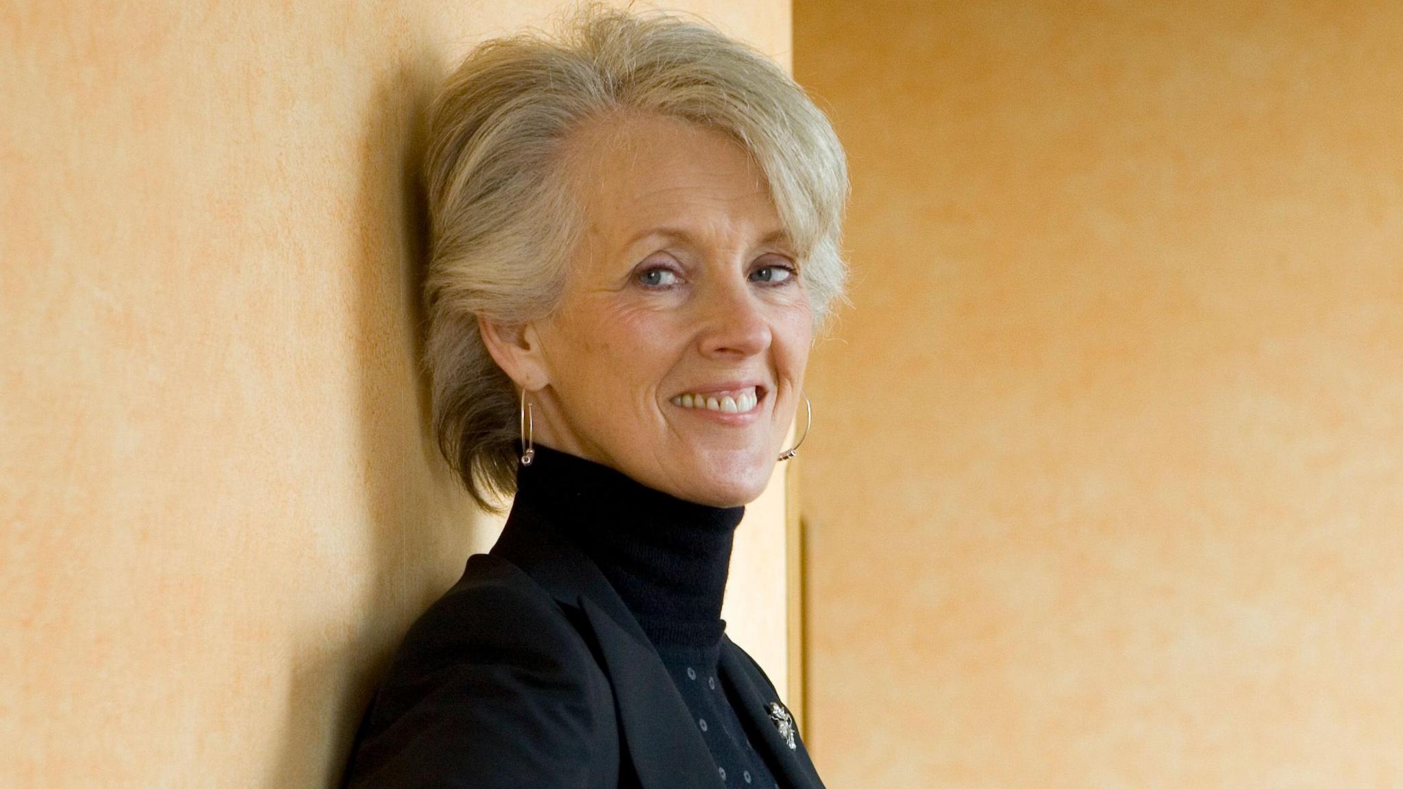 Joanna Trollope leaning against a stone well outdoors, smiling and posing for the camera.