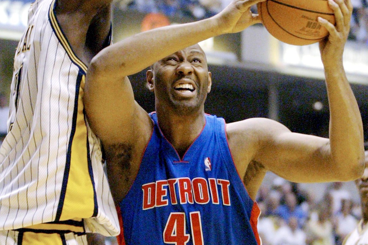 Elden Campbell in a Detroit Pistons uniform, reaching up toward the basket during an NBA game.