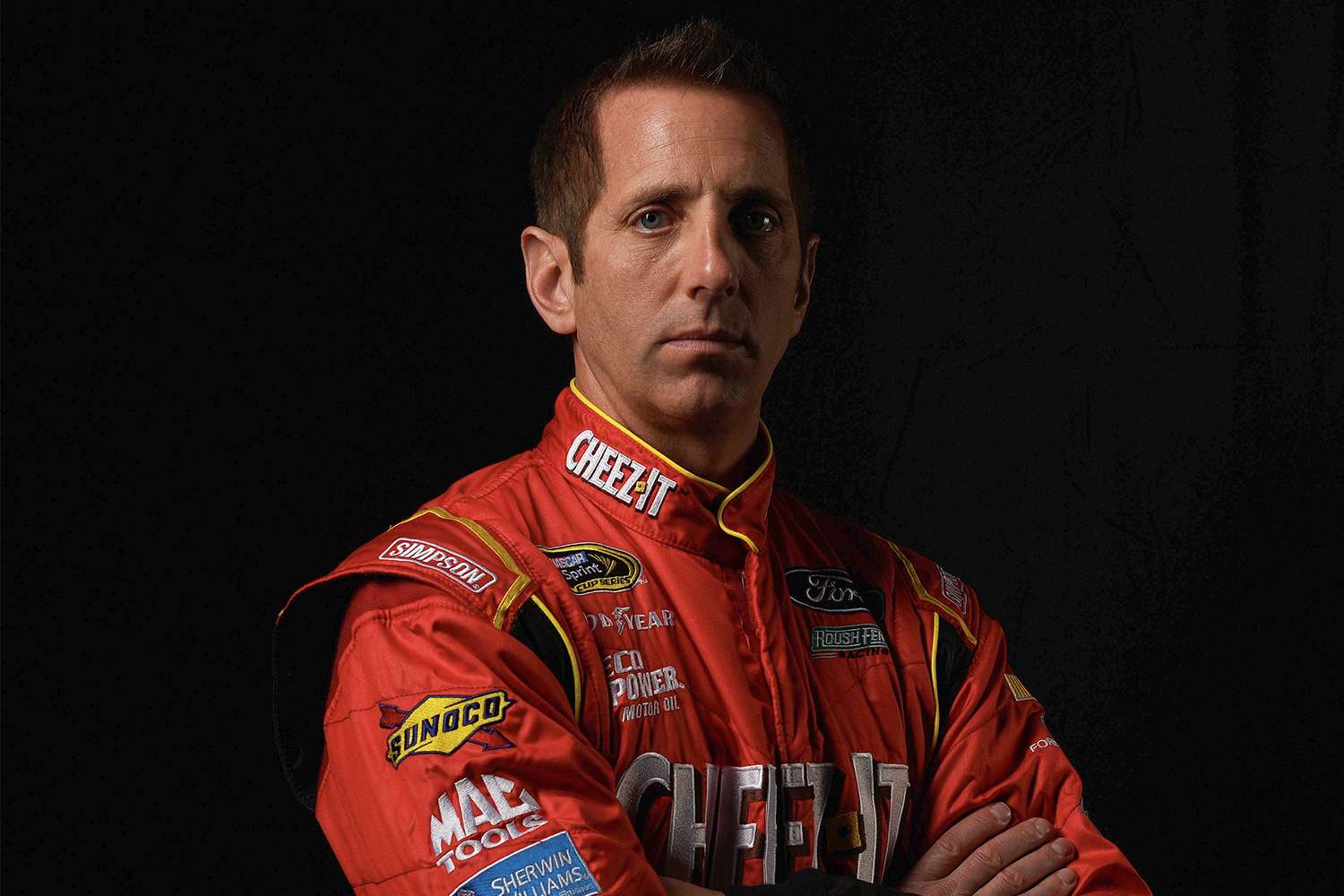 Greg Biffle posing in red NASCAR racing overalls against a dark black background.