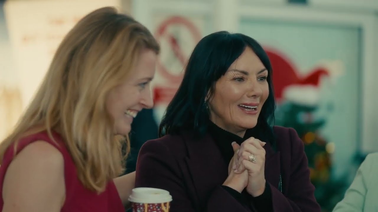 Martine McCutcheon smiling while holding a Costa Coffee cup during a festive advert at an airport arrivals hall.