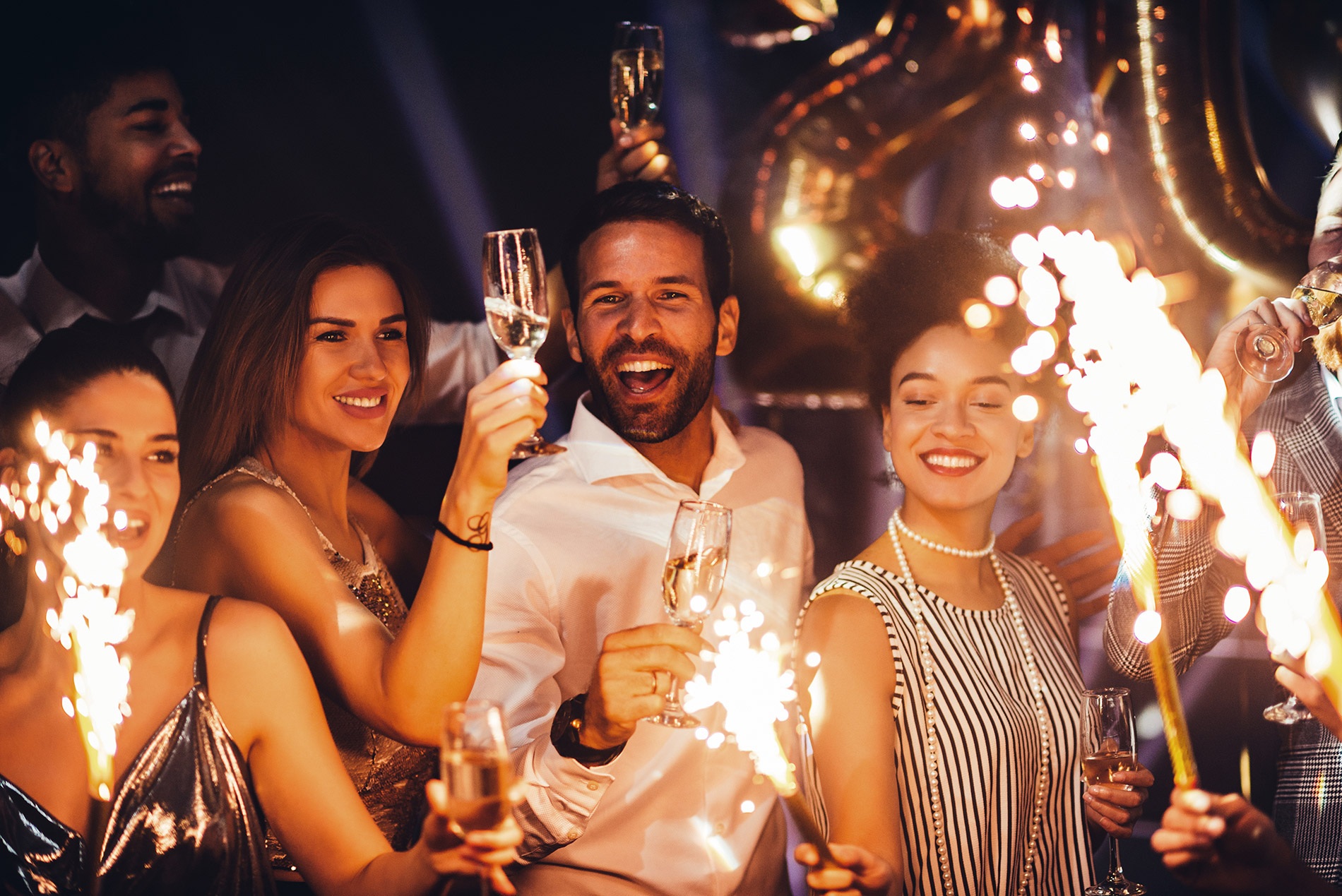 A group of friends celebrating New Year’s Eve at home, holding sparklers and surrounded by black, gold, and silver balloons.
