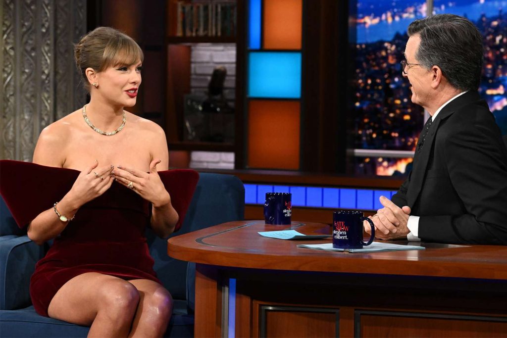 Taylor Swift sitting on The Late Show with Stephen Colbert set, talking with Stephen Colbert while wearing a burgundy velvet David Koma minidress.