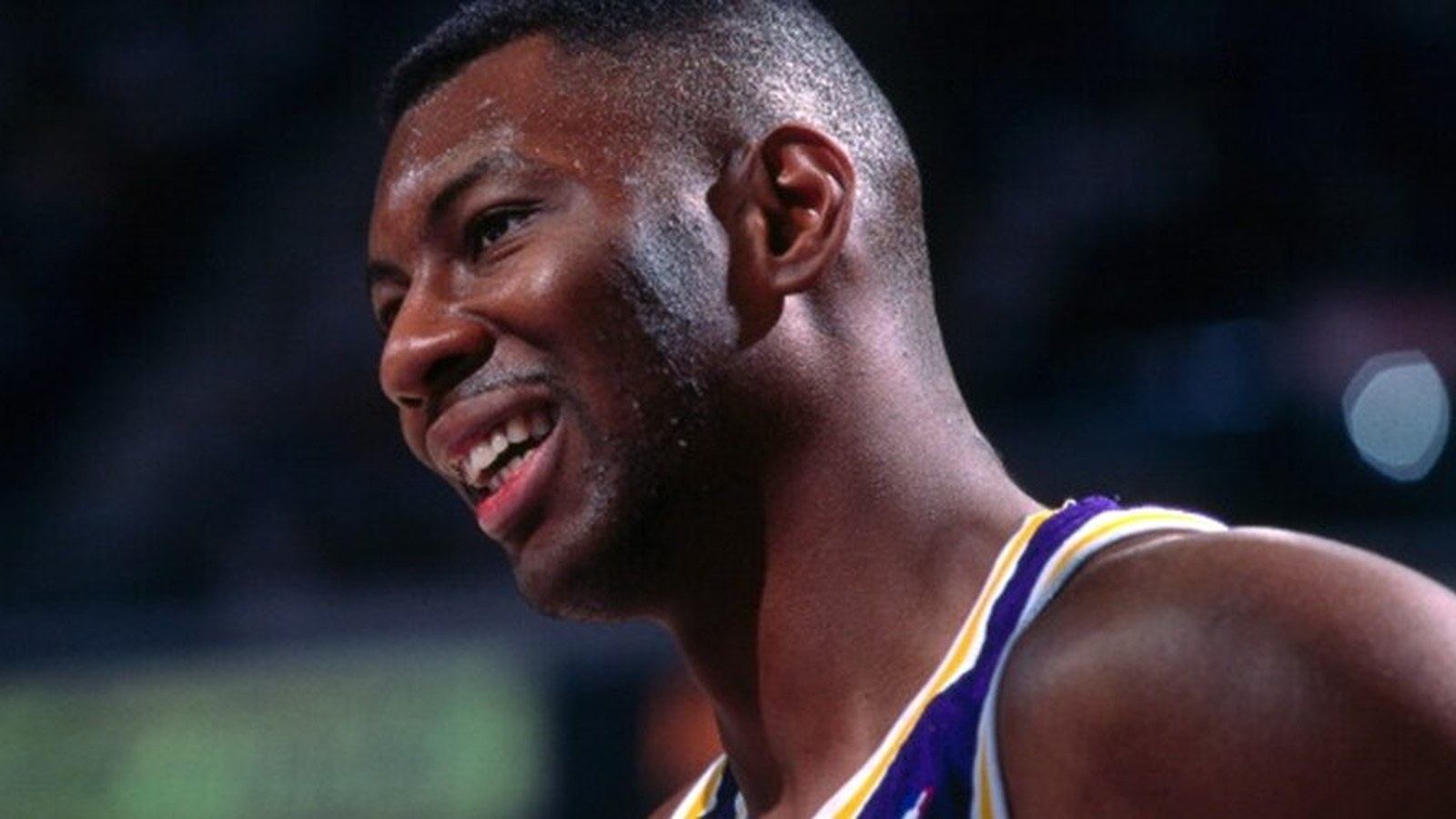 Elden Campbell wearing a Los Angeles Lakers jersey, jumping to make a shot during an NBA game, with defenders around him.