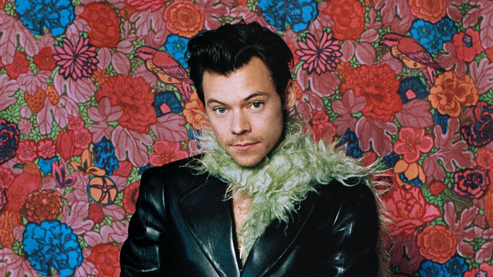 Harry Styles posing in front of a vibrant floral backdrop, wearing a stylish outfit and smiling at the camera.