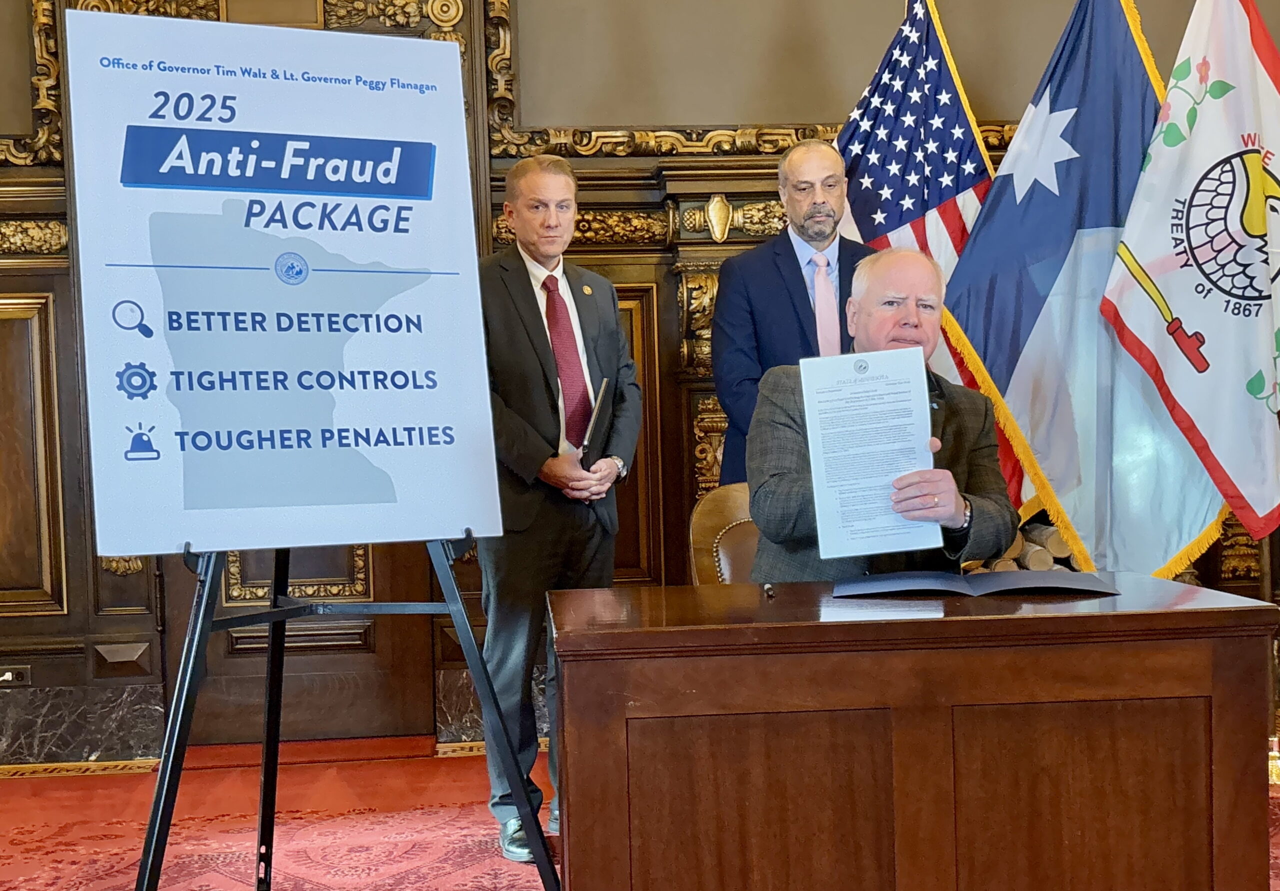 Minnesota Governor Tim Walz seated at a table with an anti-fraud sign visible behind him