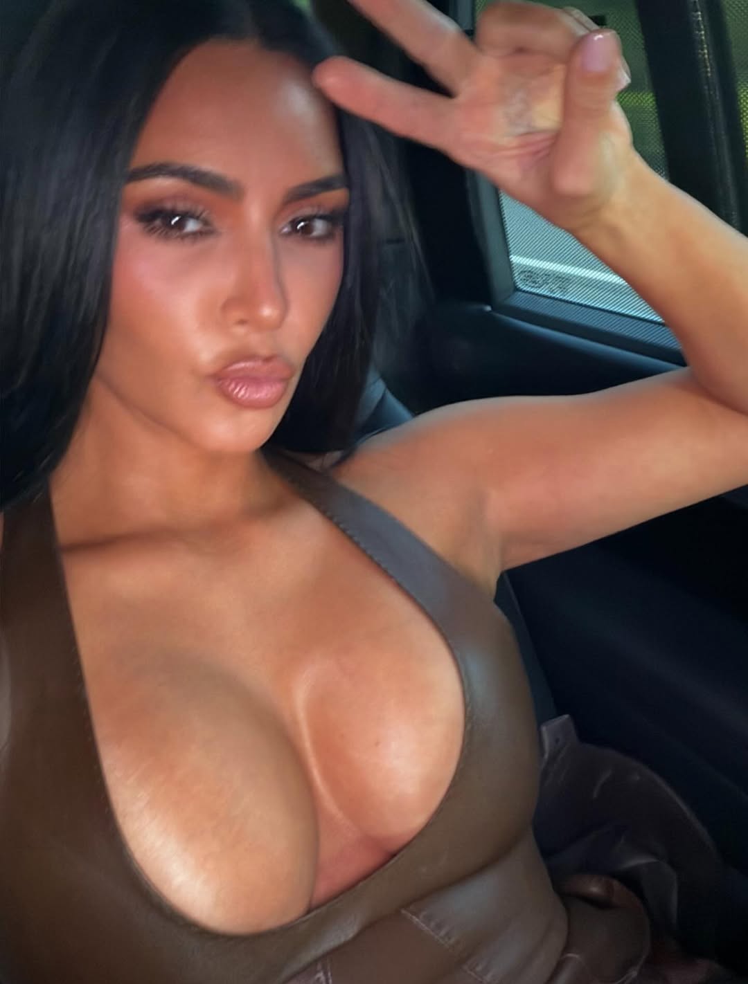 Kim Kardashian flashes a peace sign while posing in a car wearing the same plunging brown leather halter dress, blending playful attitude with high-glam fashion.