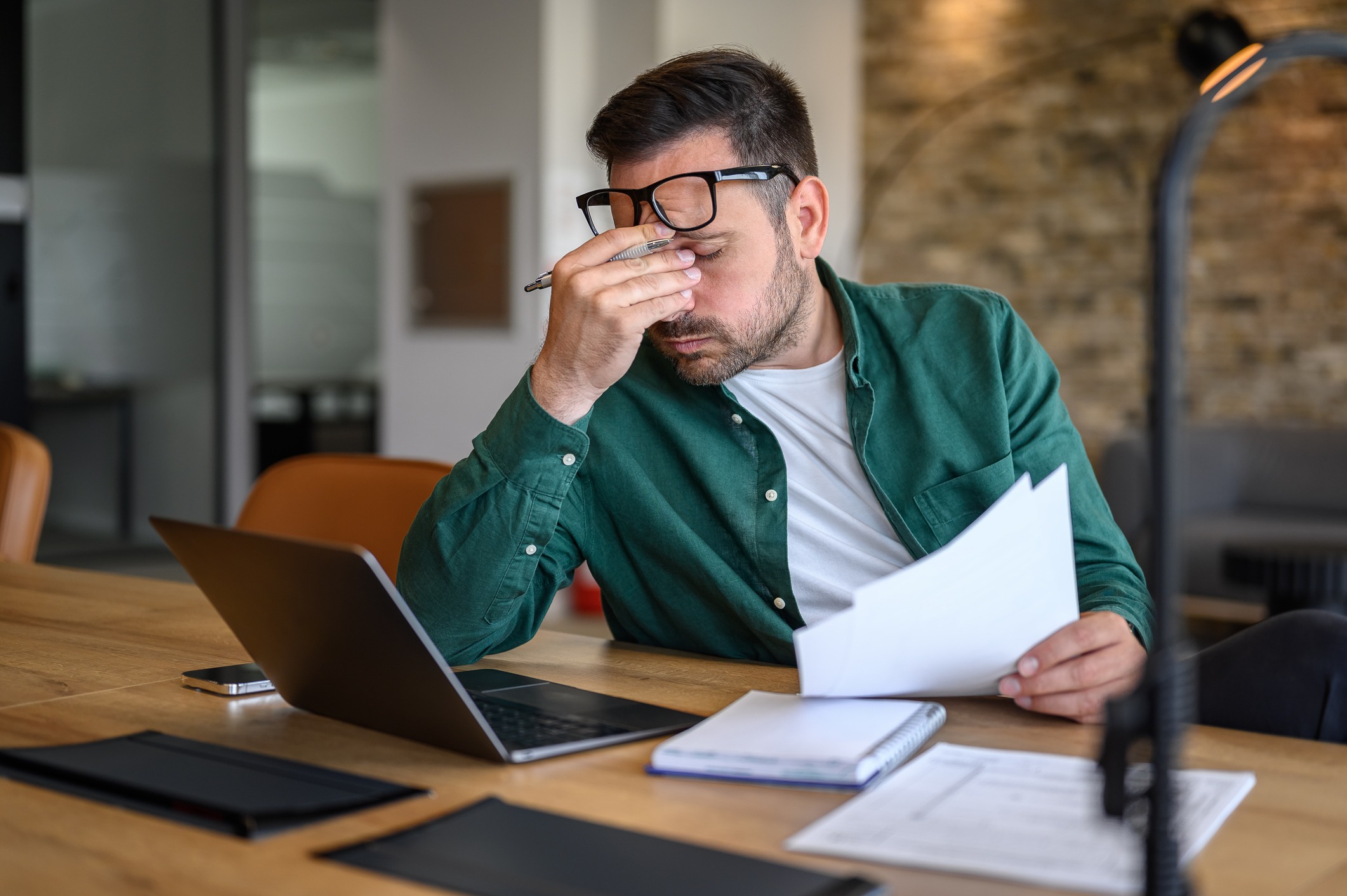 anxious male financial advisor rubbing eyes while working over laptop and analyzing reports at desk