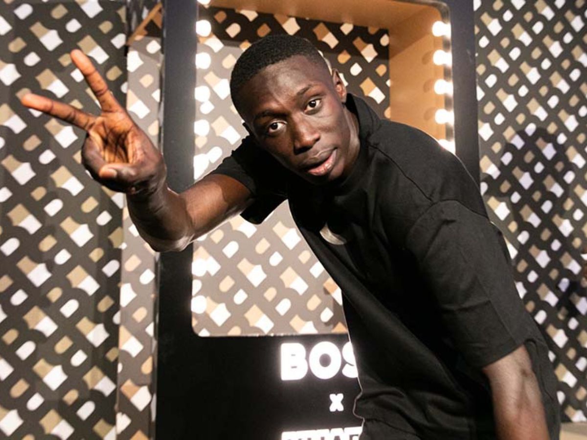 Khaby Lame on a photoshoot set smiling and flashing a peace sign at the camera