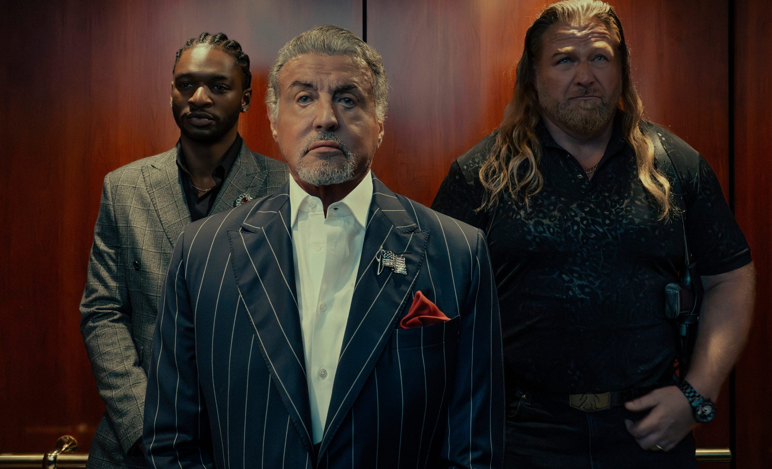 “Sylvester Stallone as Dwight ‘The General’ Manfredi standing with Big Foot and Tyson behind him in Tulsa King, dressed in a sharp suit