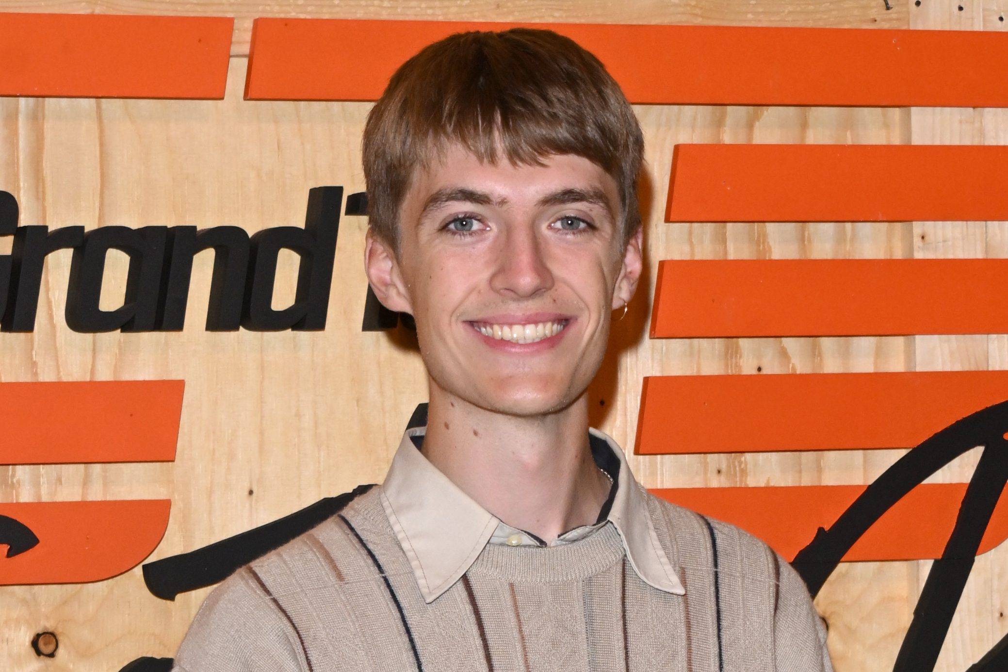 Francis Bourgeois stands in front of The Grand Tour logo smiling at the camera as the new host of the Amazon Prime Video motoring series
