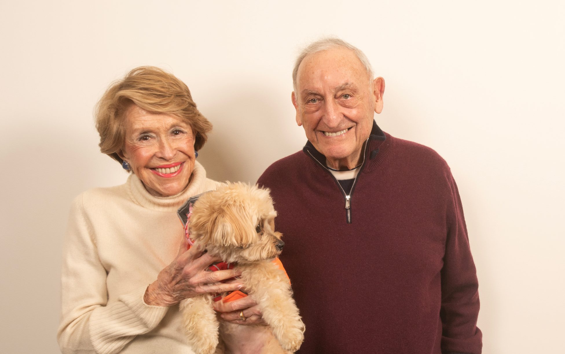 Joan and Sandy Weill stand together holding their dog Sweet Pea in a studio portrait.