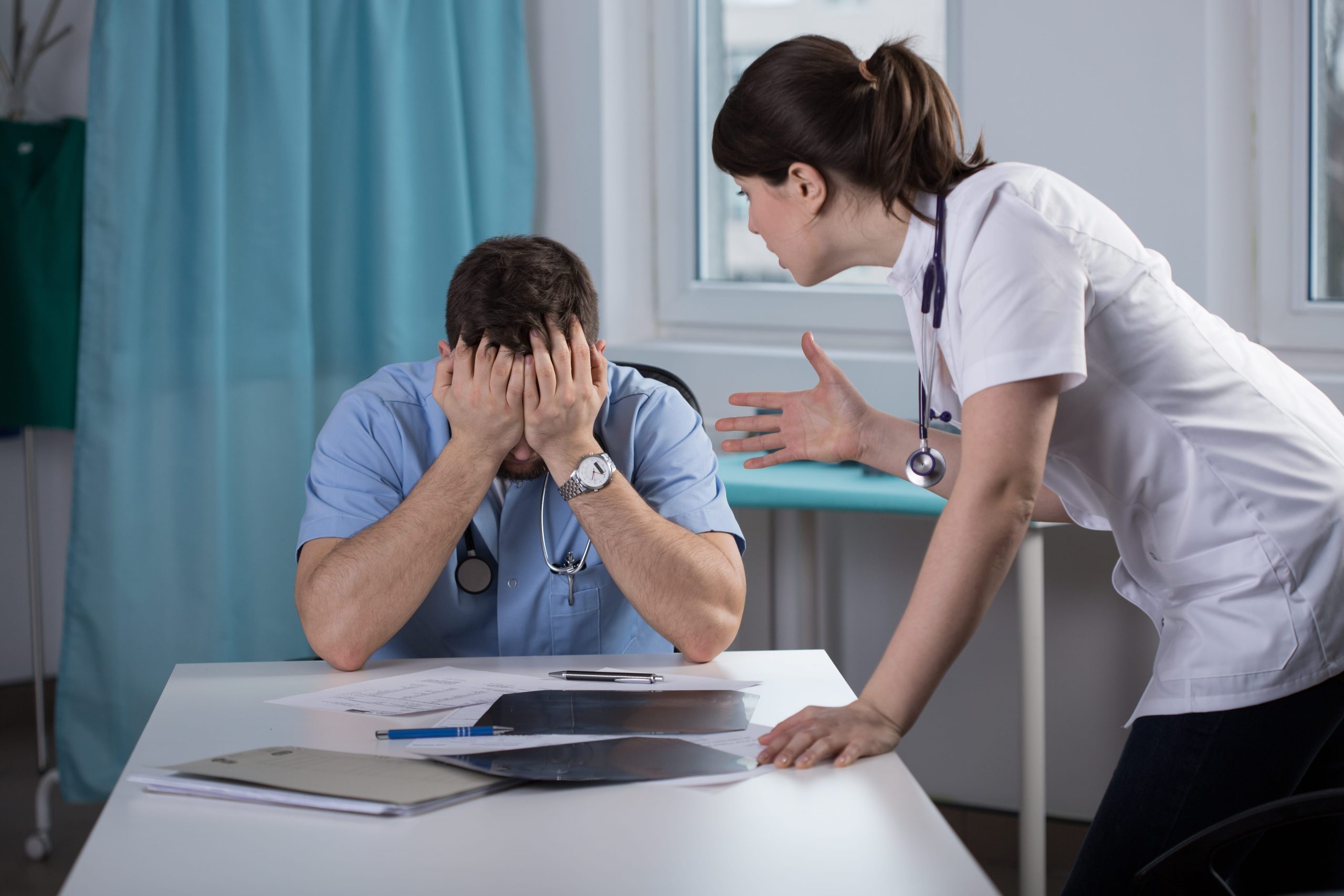 Hospital staff in a consultation room, with a clinician seated at a desk appearing distressed while a colleague speaks, highlighting professional pressure and workplace strain.