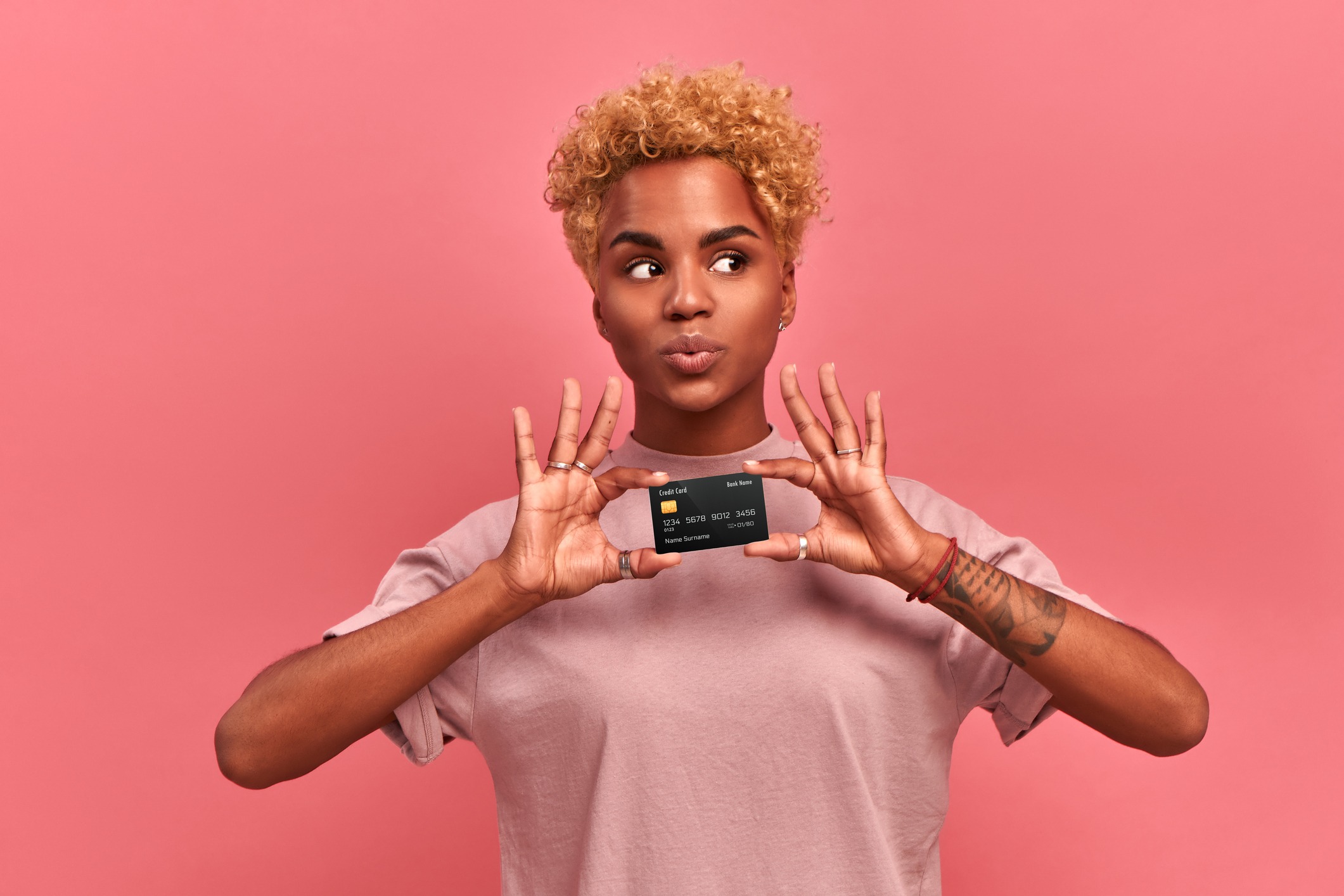 portrait of a beautiful young african american woman with blond curly hair wearing lilac clothes holding a credit card with clipping path, looks away, thinking about purchases, isolated over pink wall