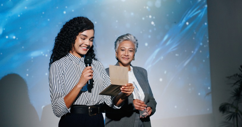 announcement, awards ceremony and presentation with business women on stage for corporate event. microphone, prize giving and reading winner with employee people in auditorium or convention center