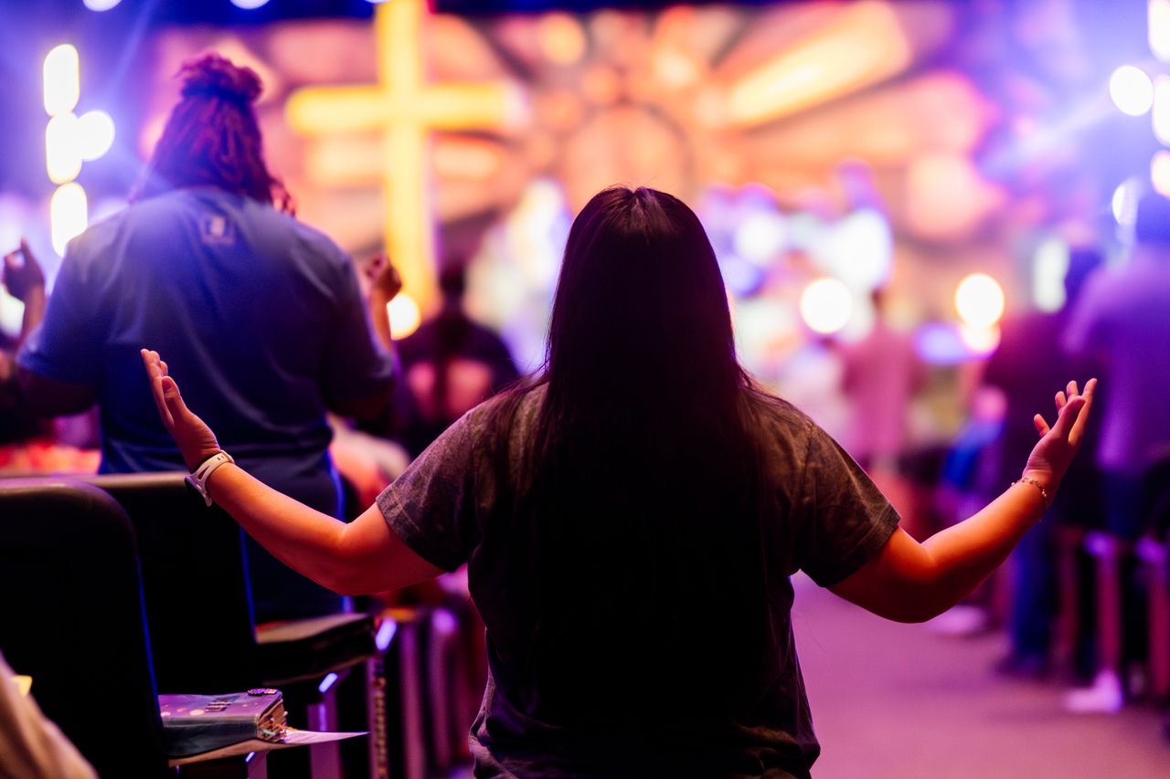 Back view of people engaged in a vibrant religious service with colorful lighting and a cross.